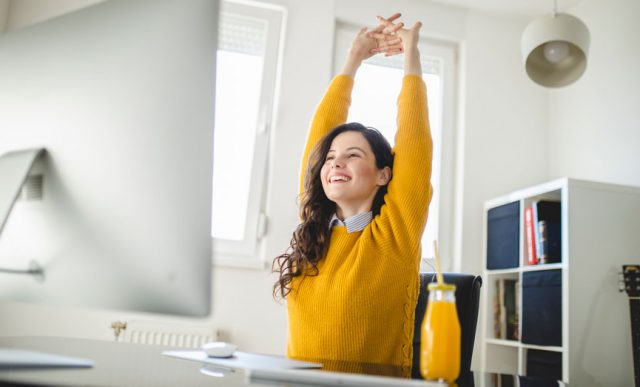 Imagem de uma mulher branca, sentada na frente de seu computador de trabalho se alongando para cuidar da saúde e bem-estar no ambiente de trabalho.