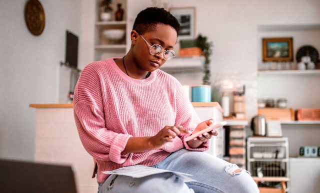 Jovem sentada com o celular na mão e com o computador enquanto analisa informações.